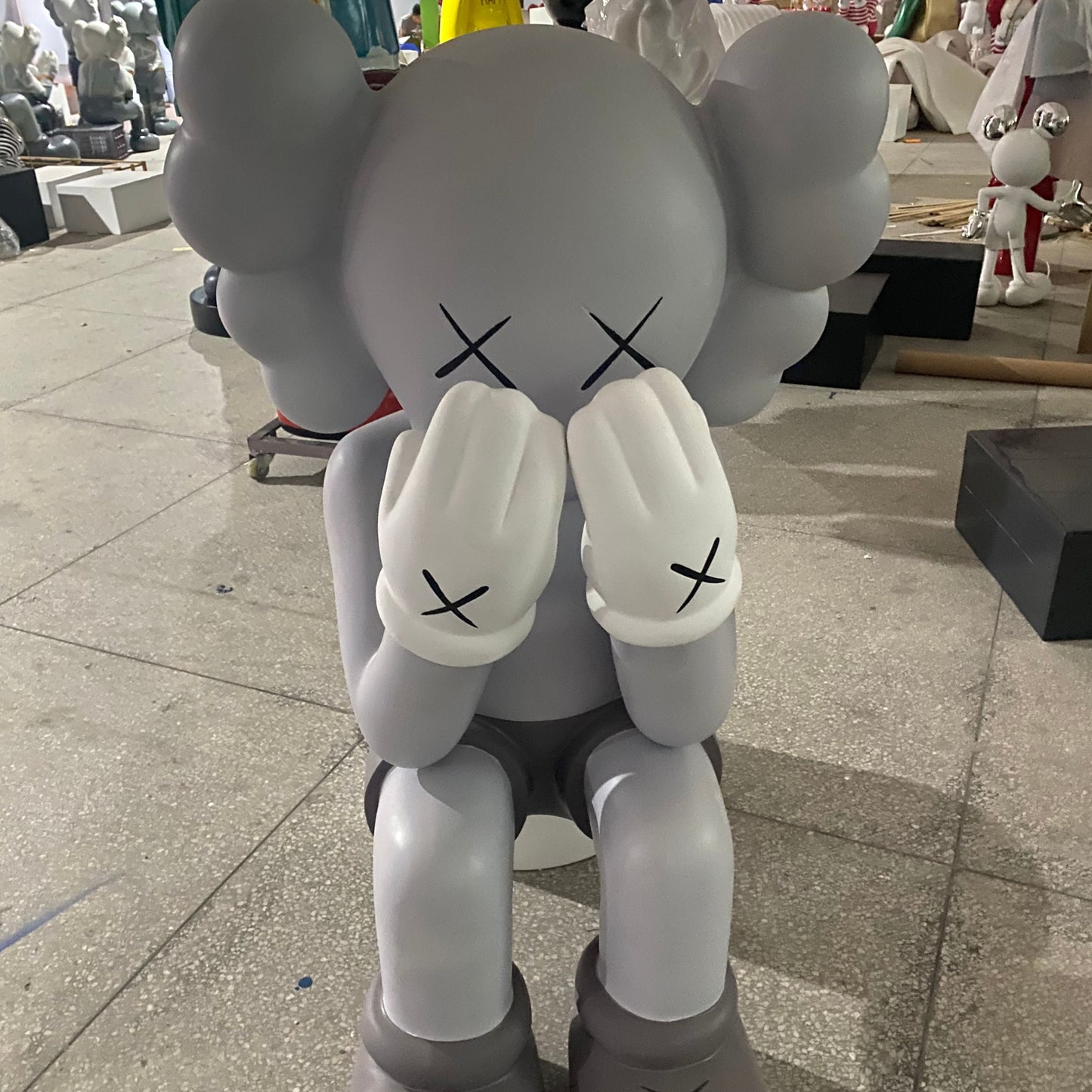 Gray cartoon-like figure with black X's on face and body in an indoor setting. kaws