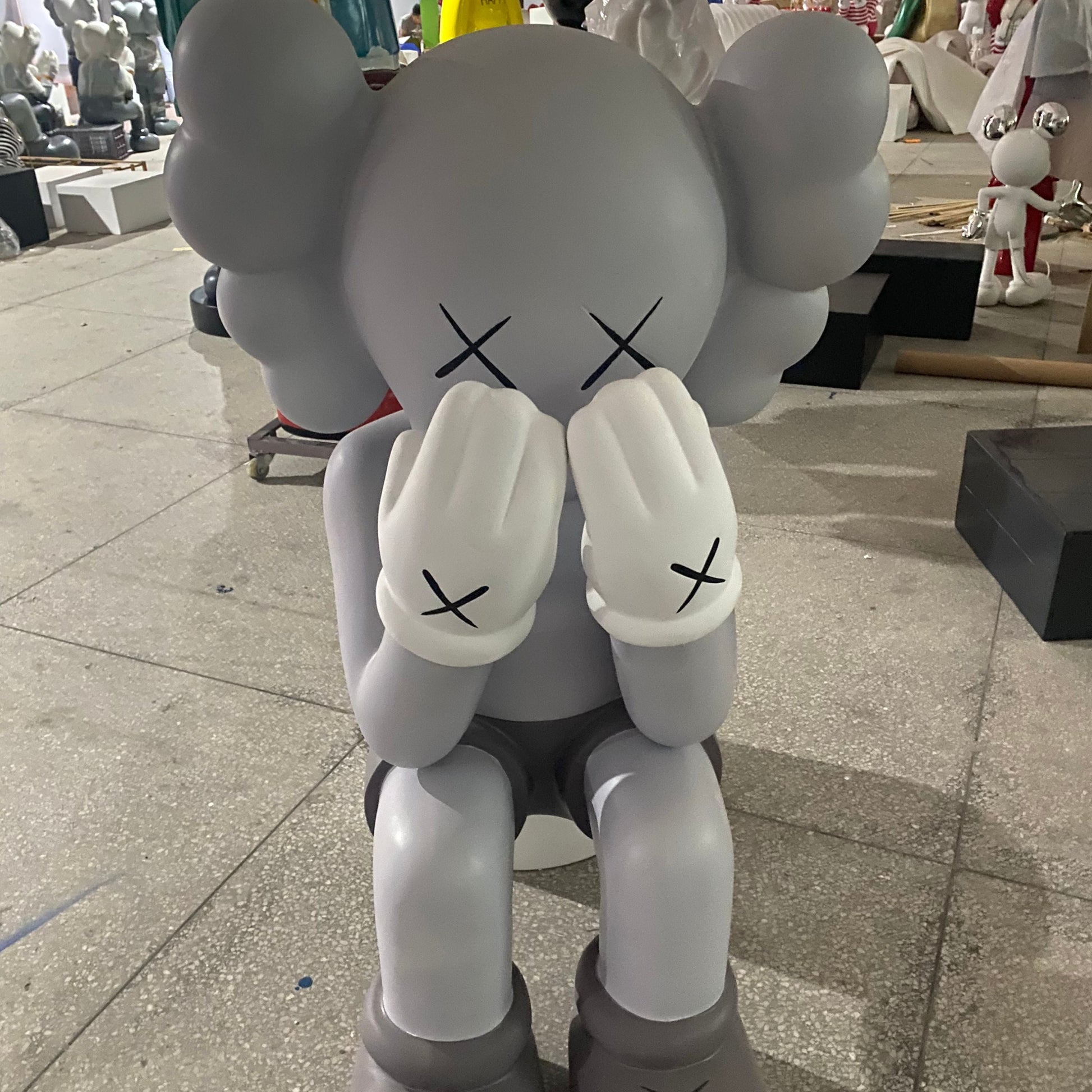 Gray cartoon-like figure with black X's on face and body in an indoor setting. kaws